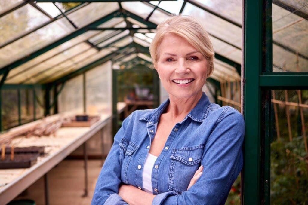 A woman stands with crossed arms in a greenhouse surrounded by lush plants