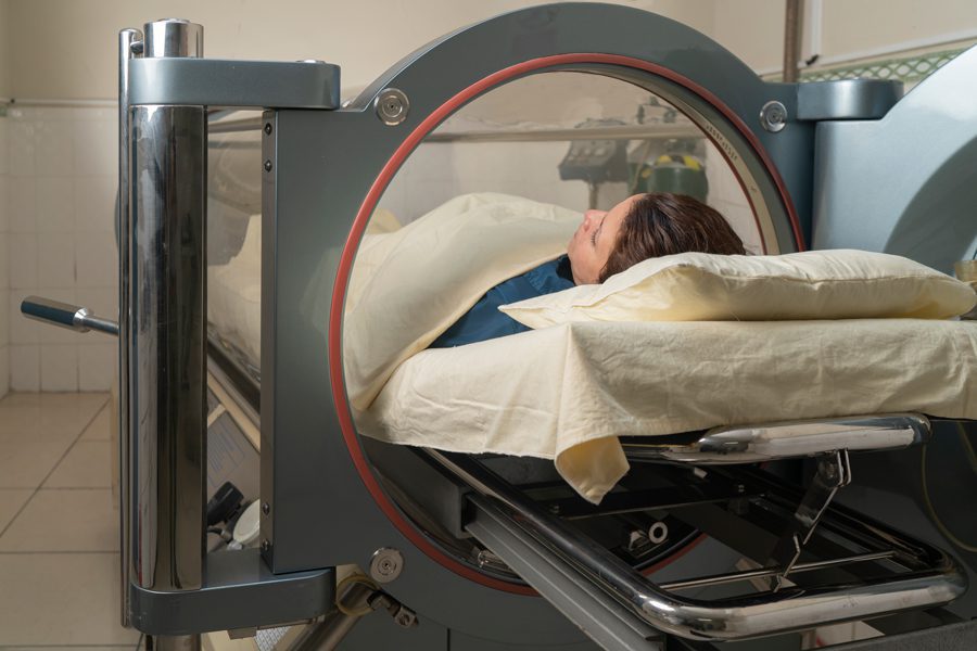 A person lies calmly in a hyperbaric chamber, partially covered with a beige blanket and resting on a pillow. The setting is clinical and serene.