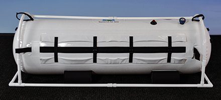 White hyperbaric chamber with a cylindrical shape, secured by black straps and a white metal frame. It features control knobs on top, conveying a clinical and functional tone.