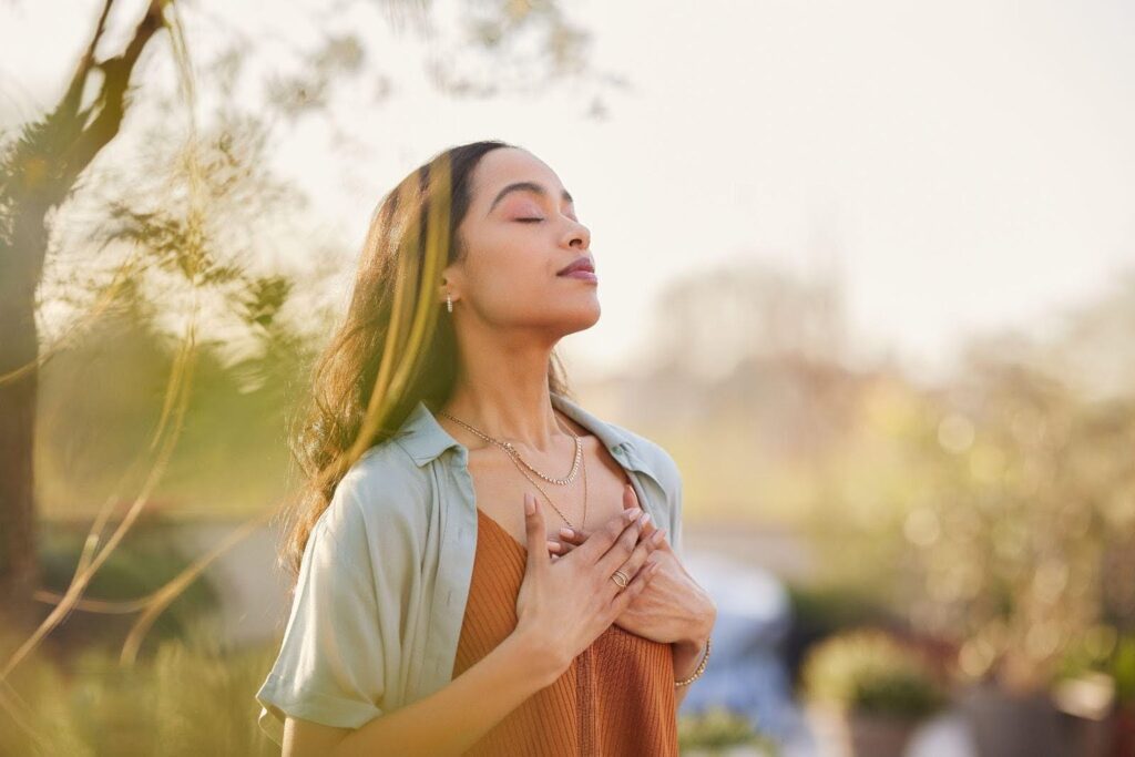 A woman stands outside taking deep breaths surrounded by nature and sunlight