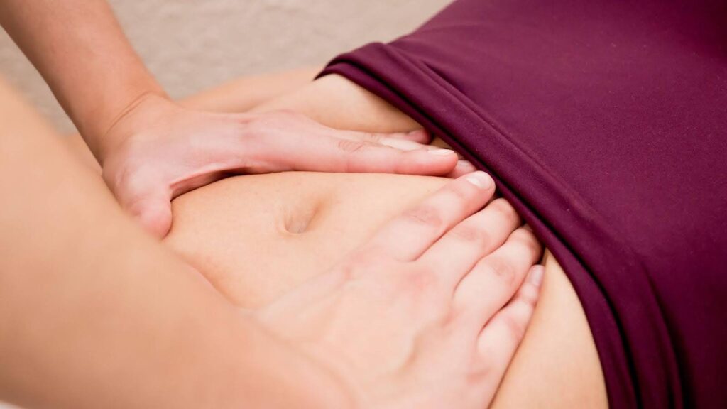A woman lies on a massage table receiving a stomach massage from a therapist in a serene setting