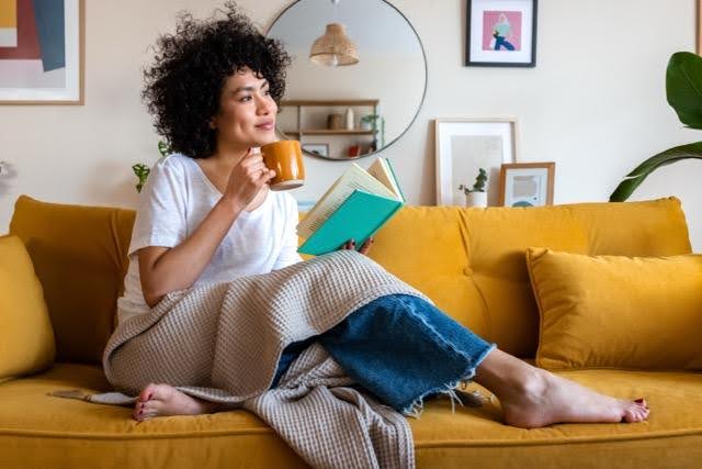 A woman with afro hair sits on a couch reading a book and sipping coffee enjoying a cozy moment