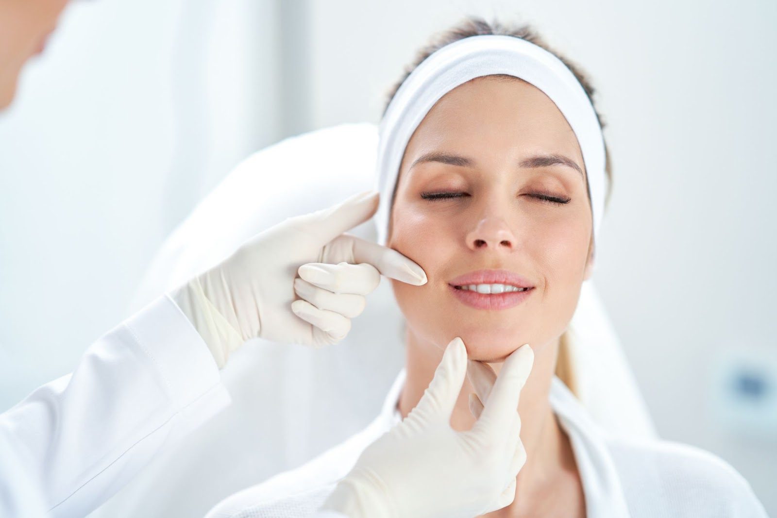 A practitioner in white gloves gently examines the face and chin of a woman with her eyes closed during a cosmetic or dermatological consultation.