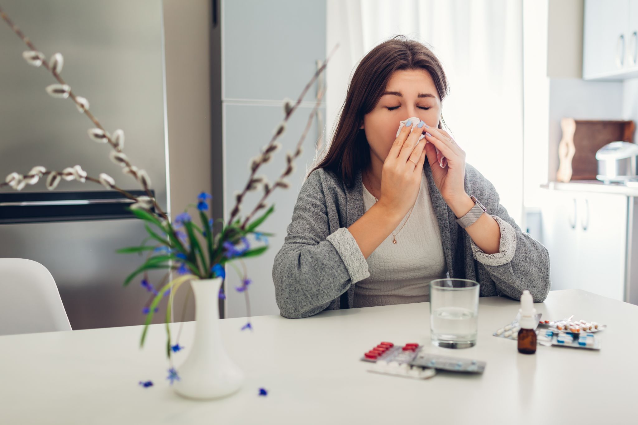 A woman blowing her nose with a tissue at a table with cold medicine and a glass of water.