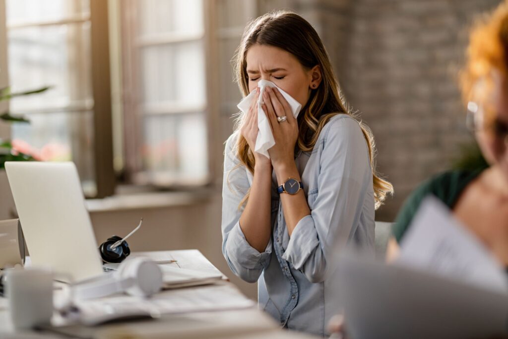 A woman sneezing or blowing her nose into a tissue while sitting at an office desk.
