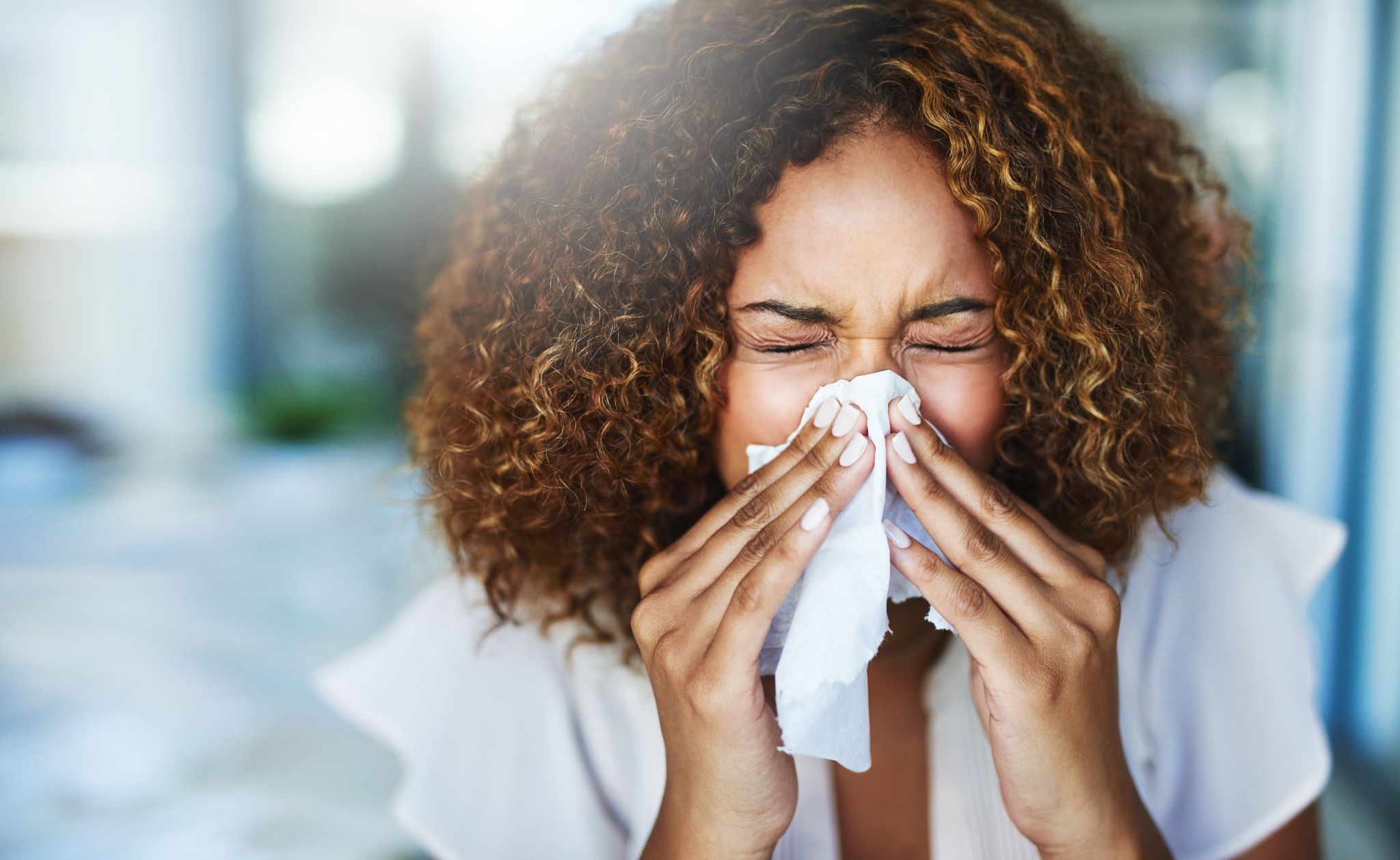 A close-up of a woman with curly hair sneezing heavily into a tissue.