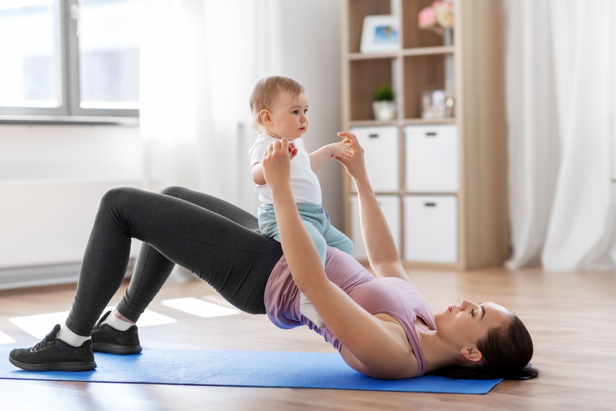 A mother performing a glute bridge exercise on a yoga mat while holding her baby on her lap.