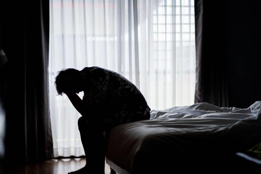 A man is seated on a bed in a dark room surrounded by shadows that enhance the mood of solitude