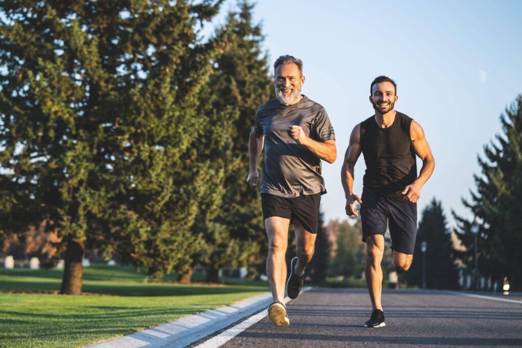 Two men jogging side by side on a paved road surrounded by greenery and a clear blue sky