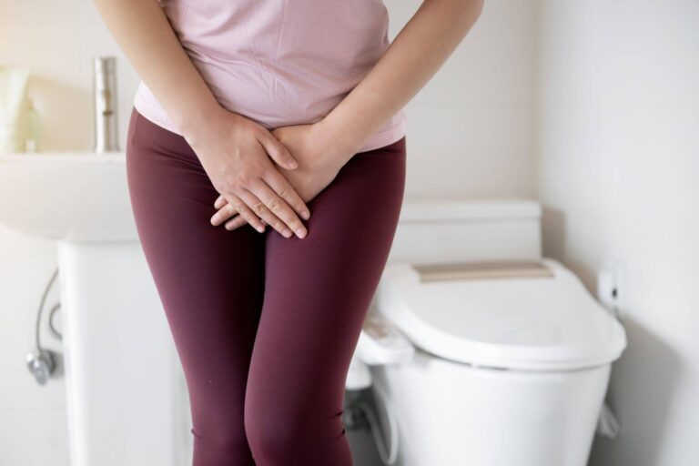 Person in pink shirt and burgundy pants stands clutching their lower abdomen before a toilet