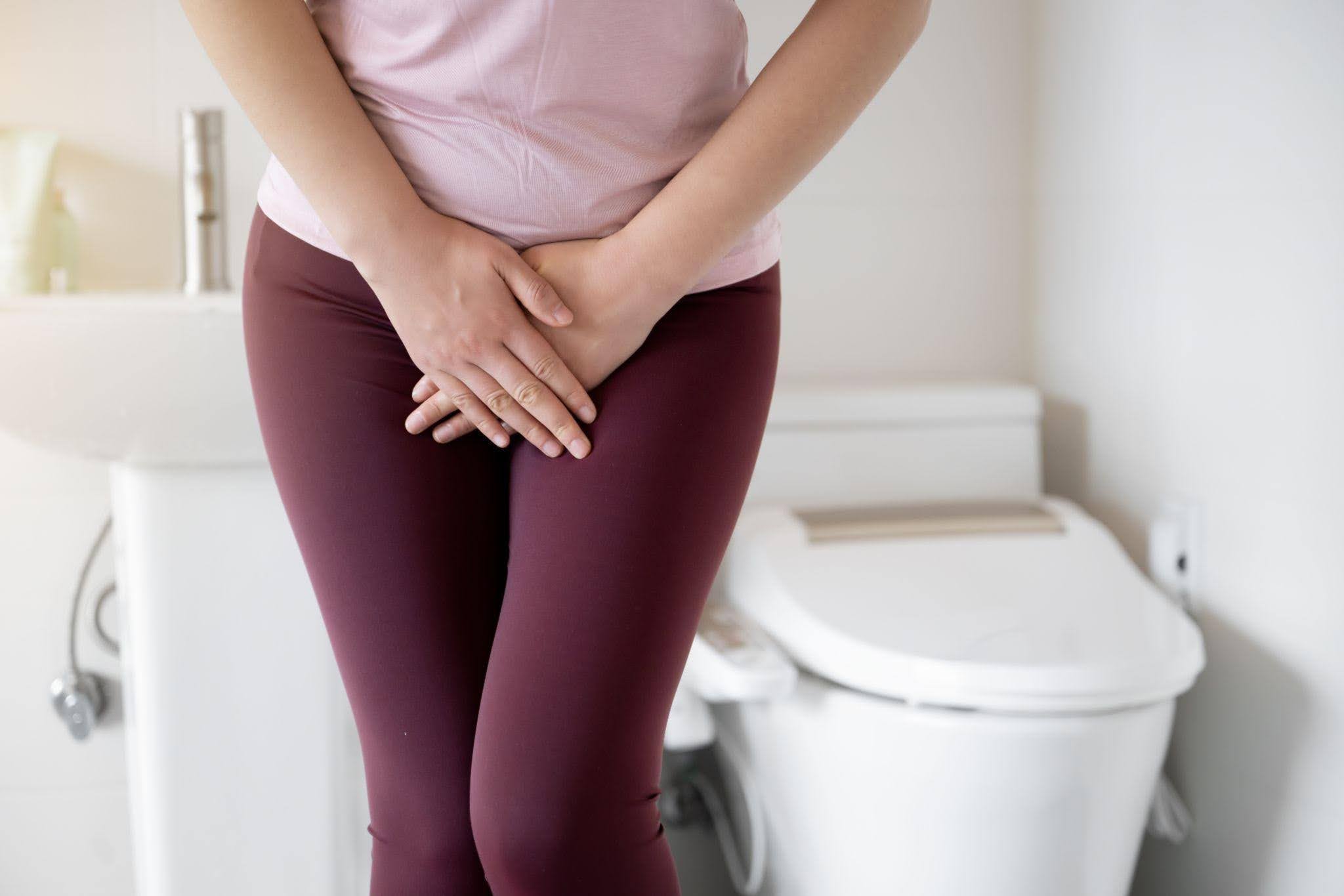 Person in pink shirt and burgundy pants stands clutching their lower abdomen before a toilet